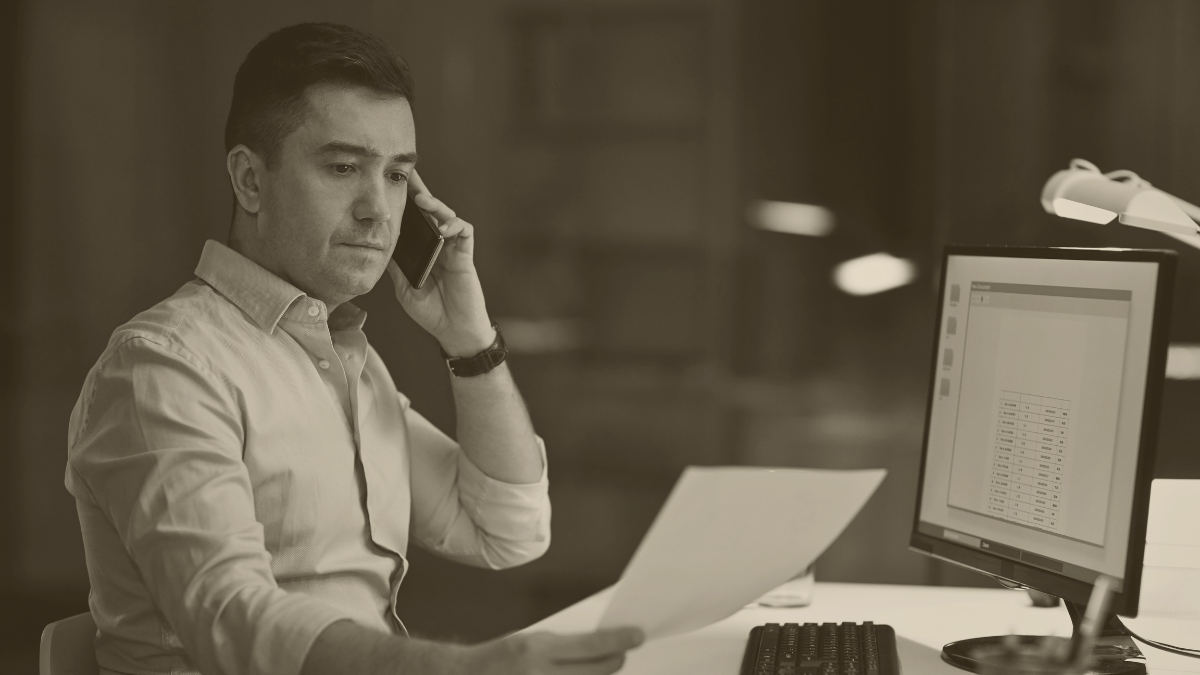 Dirigeant en chemise claire au téléphone, analysant un document devant son ordinateur dans un bureau en soirée.